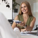 Mulher sorridente, sentada à mesa de um escritório moderno, entrega um cartão bancário a uma pessoa à sua frente, enquanto utiliza um laptop. A cena representa o atendimento personalizado oferecido pelos bancos de nicho, que se especializam em serviços financeiros segmentados, focados em atender as necessidades específicas de determinados perfis de clientes ou setores de mercado.
