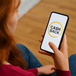 Mulher ruiva, de cabelos longos e ondulados, sentada em ambiente interno, segura um smartphone com a tela exibindo a palavra 'CASHBACK' sobre um fundo branco. A cena representa o uso de aplicativos financeiros que oferecem programas de recompensas, destacando a praticidade e os benefícios que o cashback proporciona aos consumidores nas compras e transações digitais.