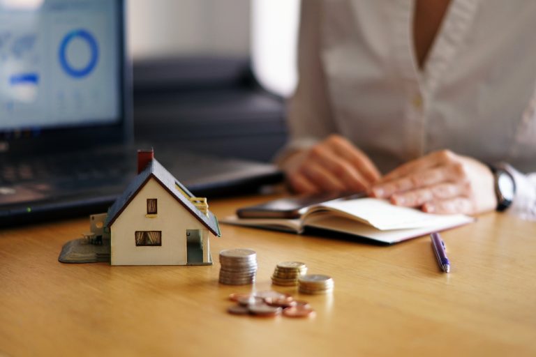 Início 8 Miniatura de uma casa posicionada sobre uma mesa, ao lado de pequenas pilhas de moedas, simbolizando planejamento financeiro. Ao fundo, uma pessoa faz anotações em um caderno, com um notebook aberto exibindo gráficos. A imagem representa o conceito de crédito imobiliário, destacando a importância do planejamento e da análise financeira na aquisição de imóveis.