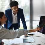 Dois profissionais analisam gráficos financeiros em um monitor de computador em um ambiente corporativo moderno, com papéis e relatórios sobre a mesa. A mulher aponta para o gráfico com uma caneta, enquanto o homem observa atentamente. A cena ilustra o estudo de tendências de mercado e a crescente importância dos ETFs no Brasil como alternativa de investimento.