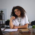 Jovem sorridente sentada à mesa de um ambiente de estudos ou coworking, com documentos, cadernos e um laptop à sua frente. Ela segura um par de óculos, transmitindo confiança e tranquilidade. A cena representa o contexto de quem busca oportunidades de financiamento estudantil, destacando o apoio financeiro que permite o acesso à educação superior e a realização de projetos acadêmicos.