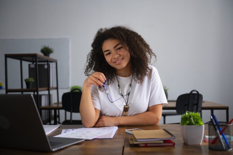 Início 16 Jovem sorridente sentada à mesa de um ambiente de estudos ou coworking, com documentos, cadernos e um laptop à sua frente. Ela segura um par de óculos, transmitindo confiança e tranquilidade. A cena representa o contexto de quem busca oportunidades de financiamento estudantil, destacando o apoio financeiro que permite o acesso à educação superior e a realização de projetos acadêmicos.