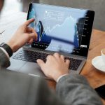 Homem de terno analisa gráficos financeiros em um laptop enquanto toma um café em uma cafeteria. Ele utiliza uma das mãos para apontar a movimentação das curvas na tela, demonstrando atenção aos detalhes. A imagem representa o processo de tomada de decisões ao investir em ações, destacando a análise de dados como parte essencial dessa atividade.