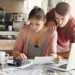 Casal jovem sentado à mesa da cozinha, analisando contas e documentos financeiros com expressão de preocupação. A mulher utiliza uma calculadora enquanto o homem observa atentamente, segurando uma caneta. A cena representa a realidade de muitas famílias que enfrentam dificuldades financeiras, destacando como os bancos estão lidando com clientes inadimplentes de forma humanizada, oferecendo suporte, negociação e soluções personalizadas para facilitar a regularização de dívidas.