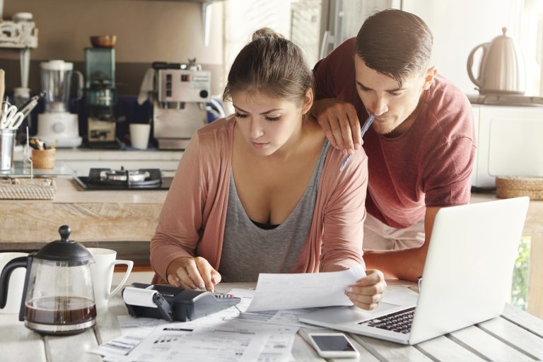 Início 24 Casal jovem sentado à mesa da cozinha, analisando contas e documentos financeiros com expressão de preocupação. A mulher utiliza uma calculadora enquanto o homem observa atentamente, segurando uma caneta. A cena representa a realidade de muitas famílias que enfrentam dificuldades financeiras, destacando como os bancos estão lidando com clientes inadimplentes de forma humanizada, oferecendo suporte, negociação e soluções personalizadas para facilitar a regularização de dívidas.