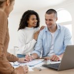 Casal feliz conversando com uma corretora de imóveis, analisando documentos sobre financiamento habitacional, com um laptop e uma maquete de casa sobre a mesa. A cena representa o processo de compra de imóvel, uma etapa comum na relação com bancos brasileiros, que oferecem diversas opções de crédito imobiliário.