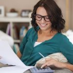 Mulher jovem e sorridente, de óculos, vestindo uma blusa verde, está sentada confortavelmente no sofá enquanto realiza cálculos financeiros com uma calculadora e analisa documentos impressos. A cena ilustra um momento de organização das finanças pessoais, representando como os bancos estão educando financeiramente seus clientes, promovendo autonomia e conscientização no planejamento financeiro.