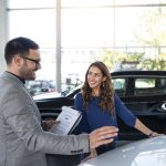 Vendedora de automóveis conversa com um cliente sorridente dentro de uma concessionária, ao lado de um carro preto. O vendedor segura um contrato enquanto explica detalhes sobre o veículo. A cena representa o processo de aquisição e negociação de financiamento para veículos, ilustrando a importância de orientações claras e soluções acessíveis na compra de automóveis.