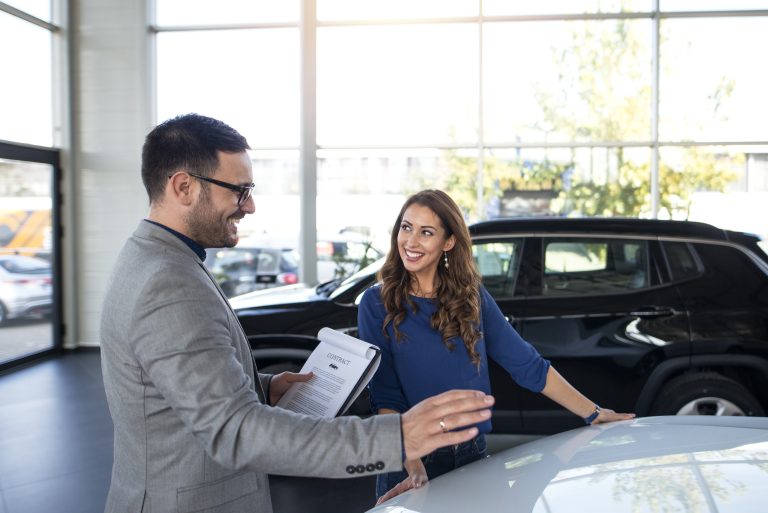 Início 6 Vendedora de automóveis conversa com um cliente sorridente dentro de uma concessionária, ao lado de um carro preto. O vendedor segura um contrato enquanto explica detalhes sobre o veículo. A cena representa o processo de aquisição e negociação de financiamento para veículos, ilustrando a importância de orientações claras e soluções acessíveis na compra de automóveis.