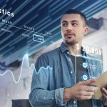 Homem segurando um tablet enquanto observa gráficos financeiros digitais e estatísticas projetadas em realidade aumentada, incluindo dados de saldo e despesas. A imagem representa o uso de tecnologias modernas por fintechs para oferecer soluções inovadoras de gestão financeira e análise de dados em tempo real.
