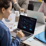Equipe de profissionais reunida em escritório, analisando gráficos e indicadores financeiros em telas de notebook e relatórios impressos. A cena destaca o uso de inteligência artificial para interpretação de dados e apoio na tomada de decisões estratégicas em ambientes corporativos.