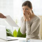 Mulher sentada à mesa de escritório, com expressão de surpresa e preocupação, cobrindo a boca com as mãos enquanto recebe um documento de uma pessoa fora do quadro. Sobre a mesa, há um laptop e relatórios financeiros. A imagem simboliza situações delicadas relacionadas ao risco de crédito, representando os impactos emocionais e profissionais causados por inadimplência, restrições financeiras ou decisões de crédito negativas.