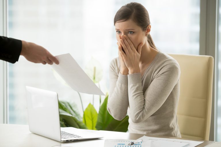 Início 10 Mulher sentada à mesa de escritório, com expressão de surpresa e preocupação, cobrindo a boca com as mãos enquanto recebe um documento de uma pessoa fora do quadro. Sobre a mesa, há um laptop e relatórios financeiros. A imagem simboliza situações delicadas relacionadas ao risco de crédito, representando os impactos emocionais e profissionais causados por inadimplência, restrições financeiras ou decisões de crédito negativas.